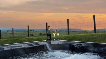 A hot tub with two glasses of white wine and a bottle, surrounded by a grassy field and a wire fence, with a sunset in the background.