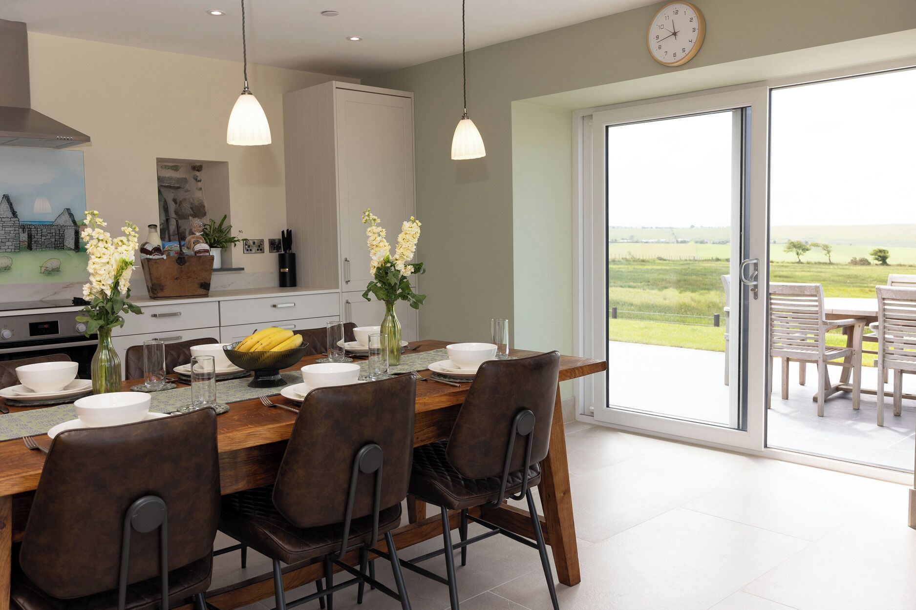 A dining area with a table set for four, a kitchen in the background, and a view of a green landscape through a sliding glass door.