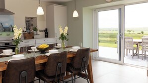 A dining area with a table set for four, a kitchen in the background, and a view of a green landscape through a sliding glass door. - Golygfa Cwrt (Talybont)