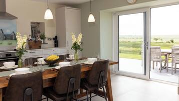 A dining area with a table set for four, a kitchen in the background, and a view of a green landscape through a sliding glass door.