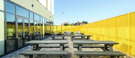 An outdoor patio with picnic tables, a yellow fence, and a building with large windows.