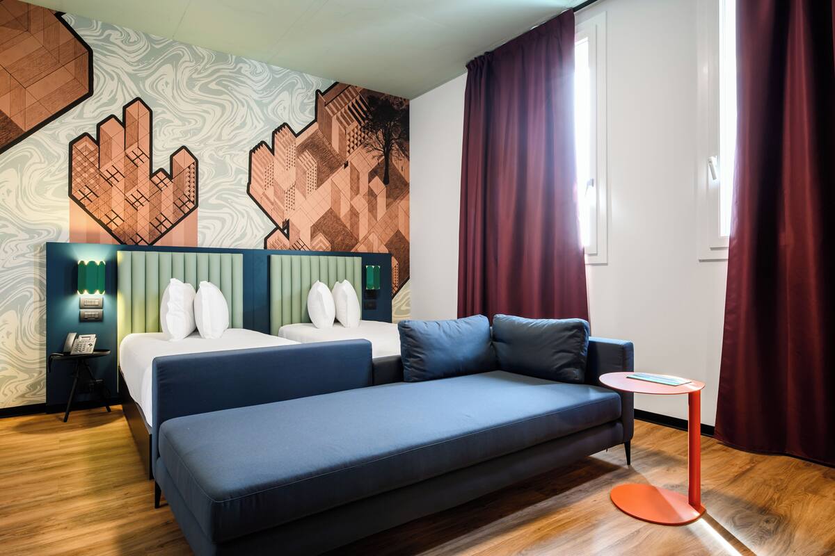 A modern hotel room with a large bed, a sofa, and a small table. The room has a geometric patterned wall and a window with curtains.