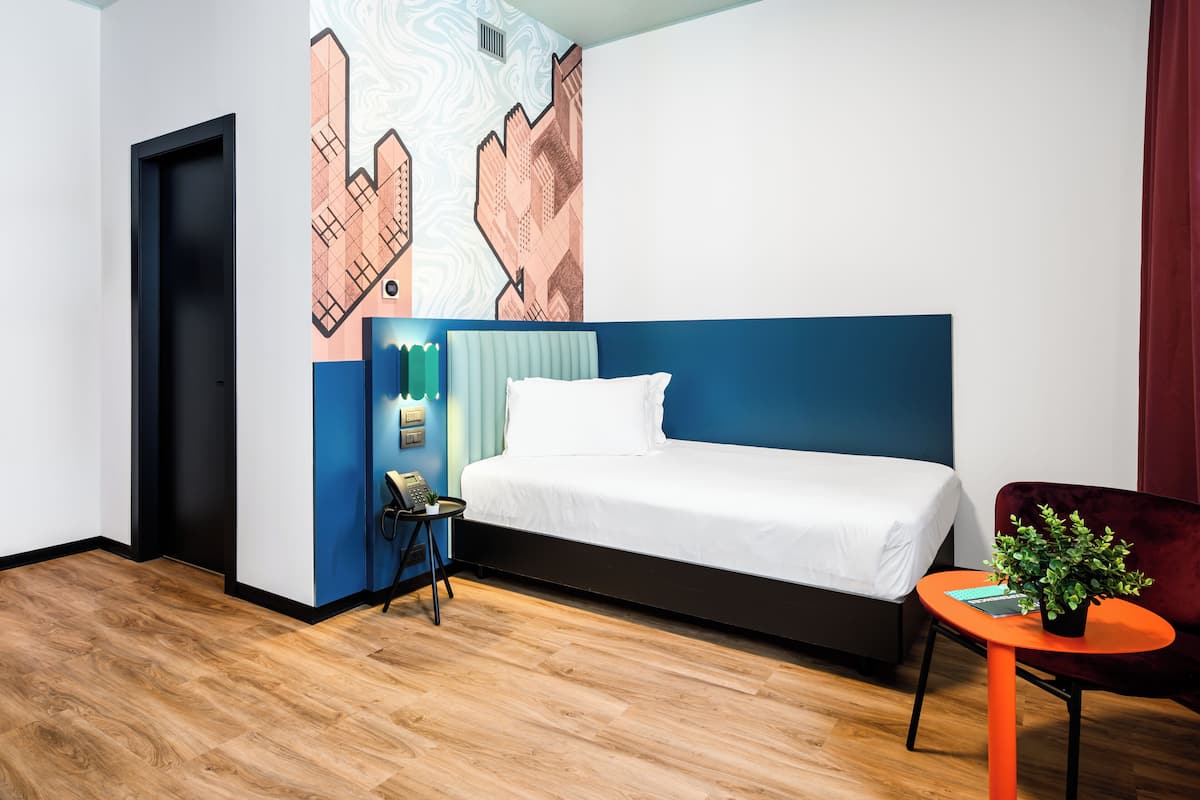 A hotel room with a bed, a small table with a plant, a chair, a wall with a geometric pattern, and a door.