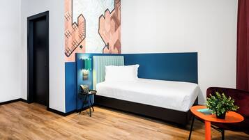 A modern hotel room with a large bed, a small table with a plant, a chair, and a wall with a geometric pattern.