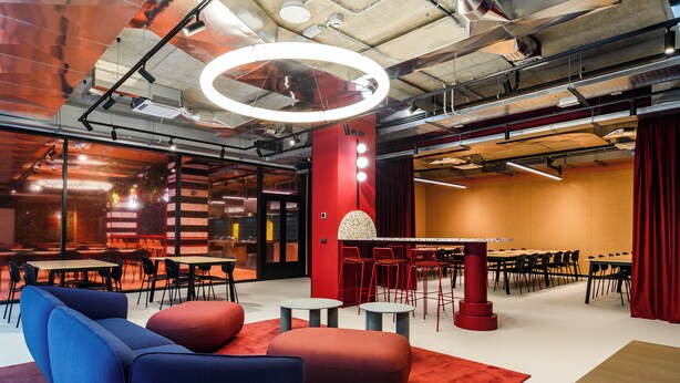A modern conference room with a red accent wall, a large circular light fixture, and a variety of seating arrangements including tables and chairs. - CX Modena Crocetta (Modena)
