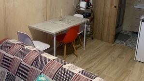 A small, cozy room with a wooden wall, a dining table with chairs, a clock, a coffee maker, and a bed with patterned bedding. - Garden studio in the Normandy countryside, near the D-Day landing beaches. (RYES)