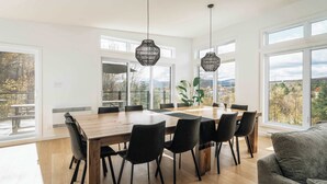 A modern dining area with a wooden table and black chairs, large windows with a view of trees and mountains, and hanging black pendant lights. - Spa Retreat with Jay Peak Views (Mansonville)