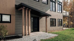 A modern two-story house with a combination of wooden and grey siding, large windows, and a covered porch area. - Spa Retreat with Jay Peak Views (Mansonville)