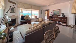Una sala de estar con sofá, sillas, chimenea, estantería y vista al mar.