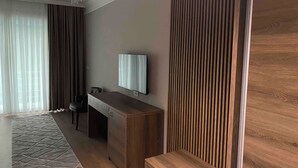 A modern hotel room with a wooden desk, a flat-screen TV, a large window with sheer curtains, and a patterned rug. - Villa Vratnica - Restaurant & Apartments (Vratnica)