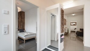 A modern hotel room with a bed, bedside table, and a mirror. There is a door leading to another room and a hallway with a light fixture.