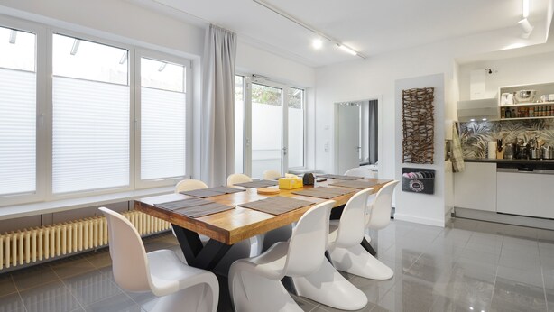 A modern dining area with a wooden table and white chairs, a kitchen with white cabinets, and large windows with blinds.