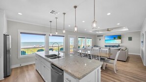 Una cocina moderna con una isla grande, muebles blancos y luces colgantes. Se ve el océano a través de amplios ventanales.