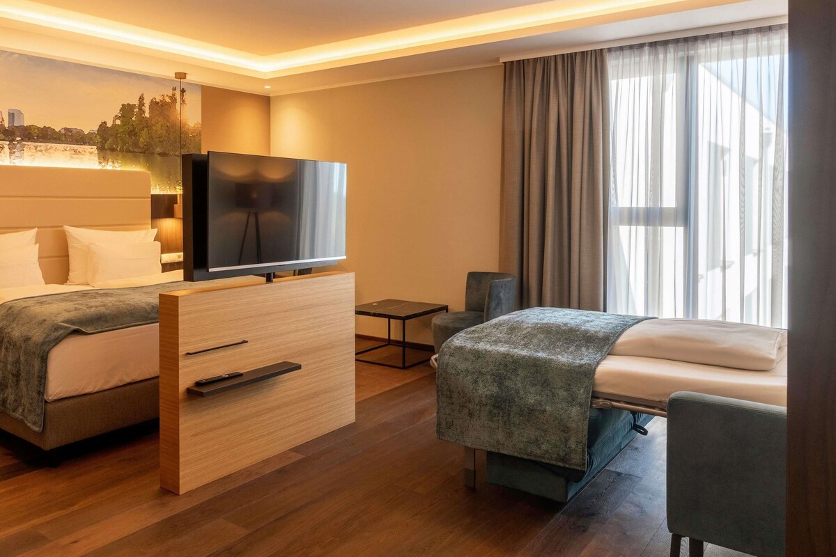 Ein Hotelzimmer mit zwei Betten, einem Fernseher, einem Schreibtisch und einem Fenster mit Vorhängen.