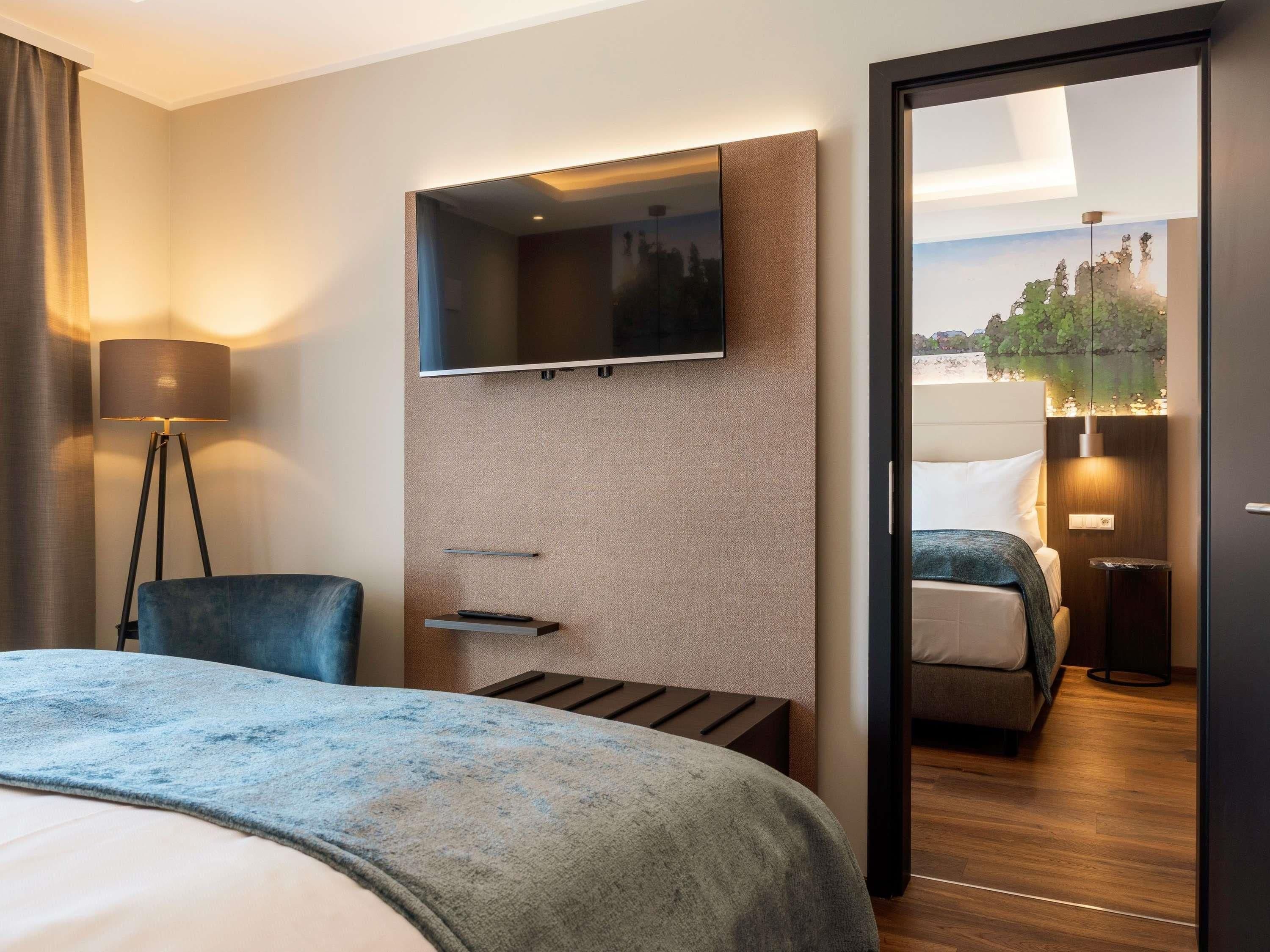 Quarto de hotel com cama, escrivaninha e cadeira, televisão de tela plana na parede e vista para a paisagem urbana através de uma porta de vidro.