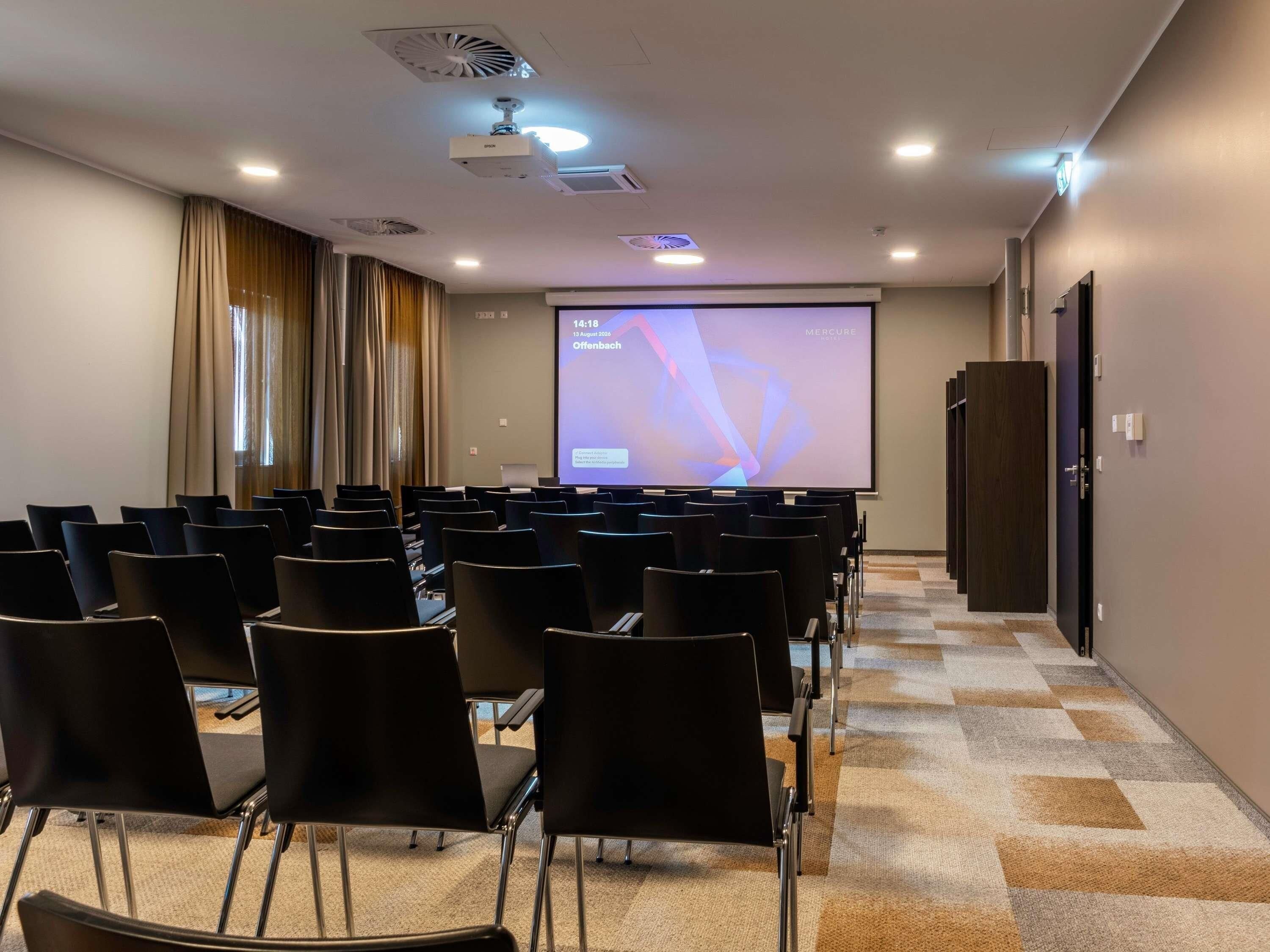 Ein Konferenzraum mit Reihen von Stühlen, die auf eine Projektionsleinwand gerichtet sind, einem an der Decke befestigten Projektor und einem Teppichboden.
