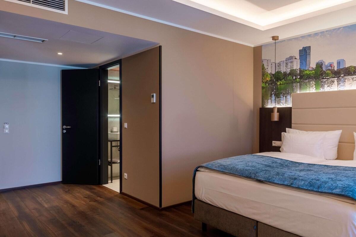 Ein Hotelzimmer mit einem Bett, einem Stadtbildgemälde, Holzboden und einer Tür zu einem Badezimmer.