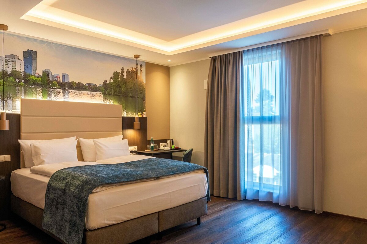 Ein Hotelzimmer mit Bett, Schreibtisch, Nachttisch, einer Stadtkulissen-Mural und einem großen Fenster mit Vorhängen.