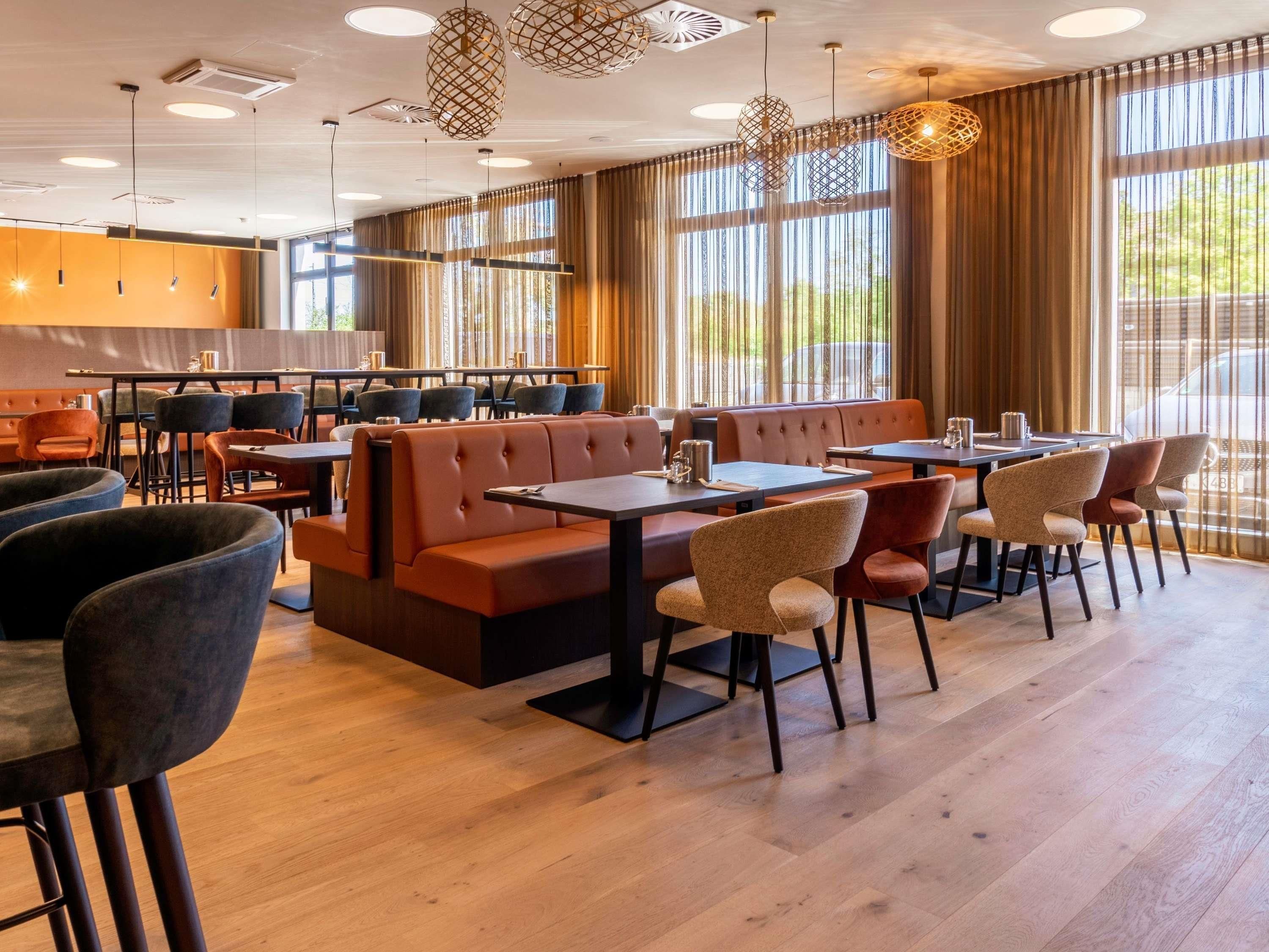 Um restaurante moderno com assentos em cabines e mesas, grandes janelas com cortinas e luminárias pendentes.