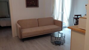 Un salon avec un canapé beige, une table basse en verre et un coin cuisine équipé d’un four à micro-ondes.