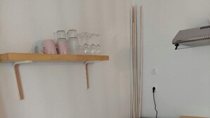 Une pièce avec une table en verre, une chaise en bois, un petit meuble, une étagère avec des verres et des tasses, et un radiateur fixé au mur.