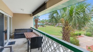A balcony with a wooden table and chairs, a glass door, and a view of a lawn and palm trees.