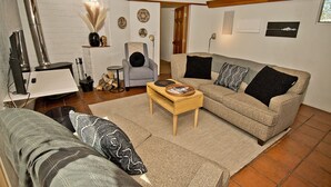 Ein Wohnzimmer mit Sofa, Sessel und Couchtisch. Ein Kamin, ein Fernseher und eine Tür zu einem weiteren Raum sind ebenfalls vorhanden.