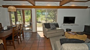 Ein Wohnzimmer mit Sofa, Fernseher, Esstisch aus Holz, Stühlen und einer Schiebeglastür, die zu einem Garten führt.