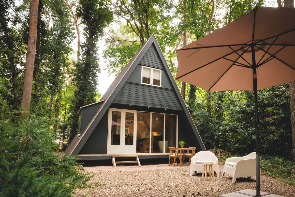 Une cabane moderne aux couleurs sombres, dotée d’un toit triangulaire, entourée d’une terrasse en gravier et de meubles en bois.