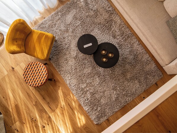 A room with a wooden floor, a grey shaggy rug, a yellow armchair, a round ottoman, and a black circular object on the rug.