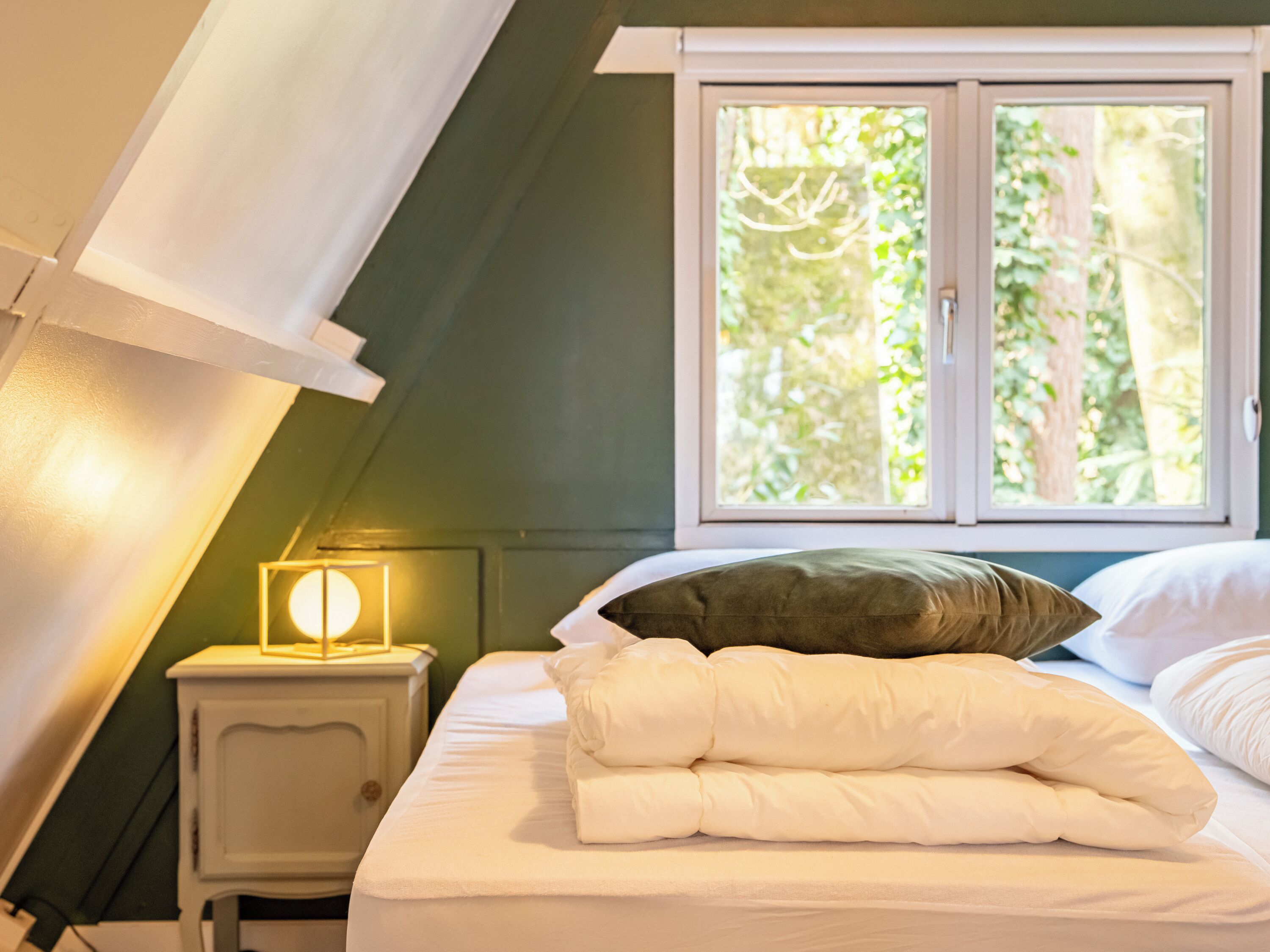 Een slaapkamer met een bed, kussens, een nachtkastje, een lamp en een raam met uitzicht op bomen.