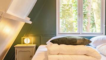 Een slaapkamer met een bed, kussens, een nachtkastje, een lamp en een raam met uitzicht op bomen.