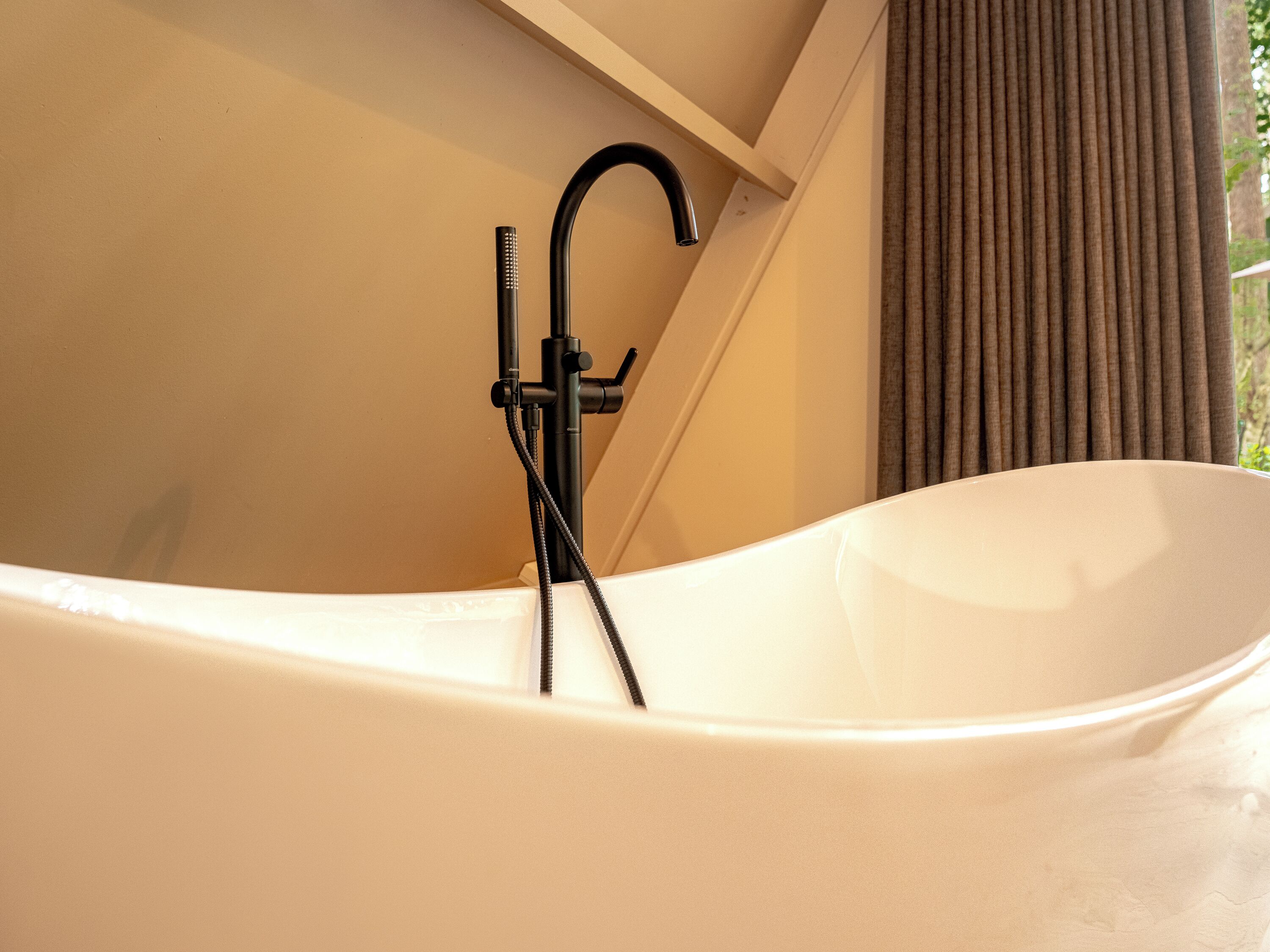 A modern bathtub with a black faucet and hose, beige walls, and a window with curtains.