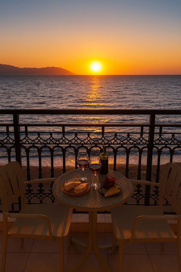 Una mesa con sillas y una botella de vino en un balcón con vistas al mar al atardecer.