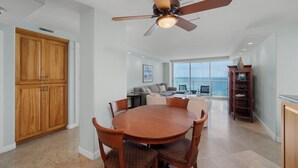 A spacious living area with a large wooden dining table, comfortable seating, a ceiling fan, and a view of the ocean through large windows.