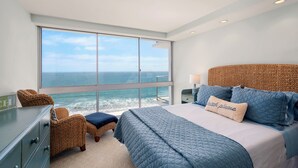 A bedroom with a large window overlooking the ocean, a bed with blue bedding, a wicker chair, and a dresser.