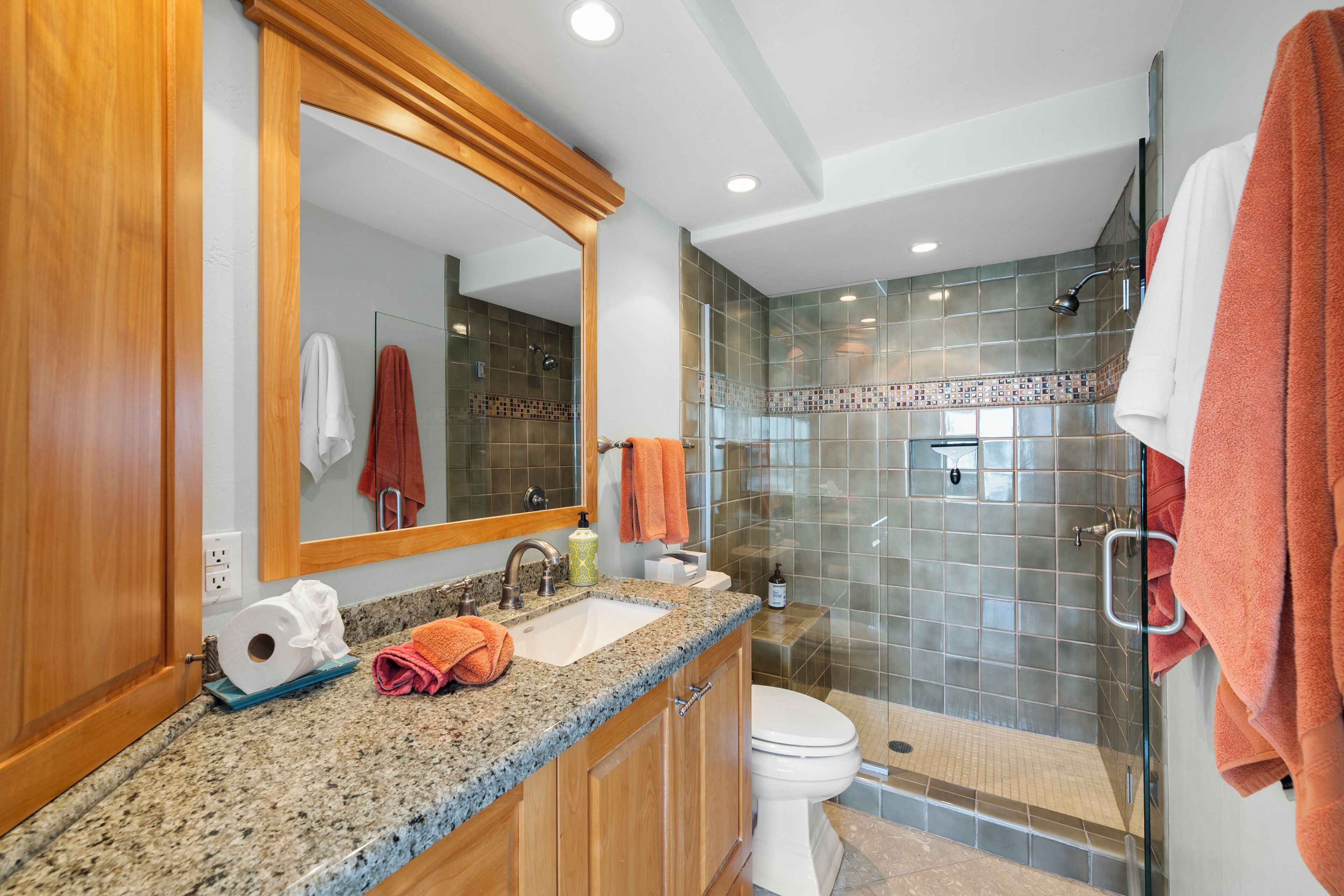 A modern bathroom with a granite countertop, wooden cabinets, a glass shower enclosure, and a white toilet.
