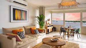 A modern living room with a sofa, coffee table, and dining area. There is a large window with a view of the sea.