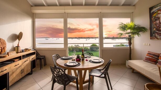 A dining area with a table set for four, large windows with a view of the sea, a ceiling fan, and a sofa with cushions.