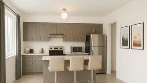 A modern kitchen with grey cabinets, a stainless steel refrigerator, a microwave, and a coffee maker. There are three bar stools at the counter. - Wasaga Luxury Coastal Escape (Wasaga Beach)