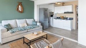 Ein modernes Wohnzimmer mit einer Couch, einem Couchtisch und einer Küchenzeile mit weißen Schränken und einem Kühlschrank.