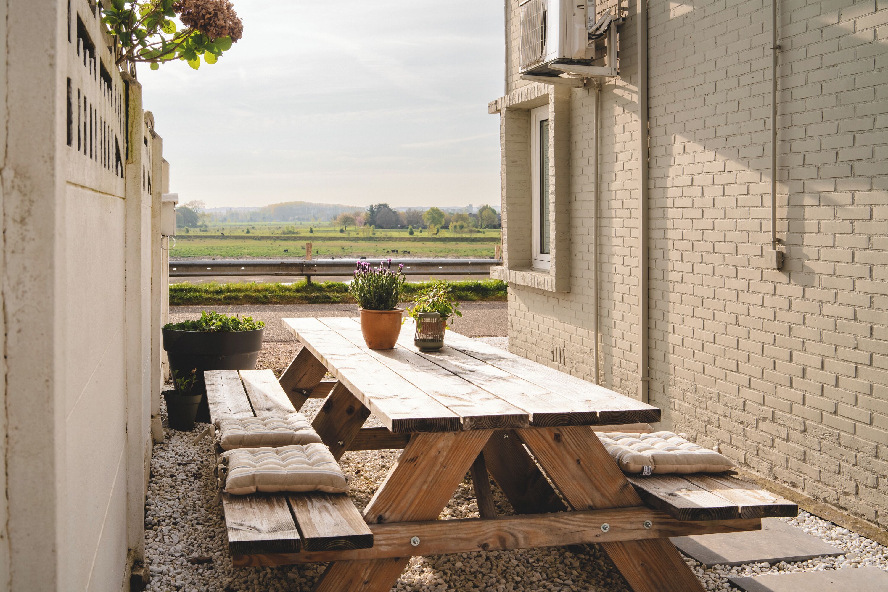 An outdoor patio with a wooden table and benches, surrounded by potted plants and a view of a green field.