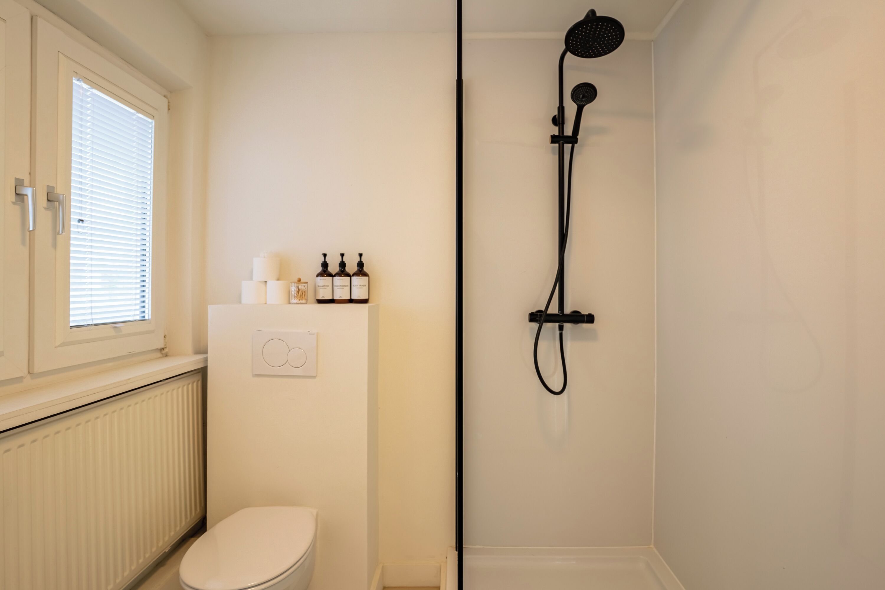Un bagno con il water, una finestra con persiane, un termosifone e una doccia con un tubo nero.