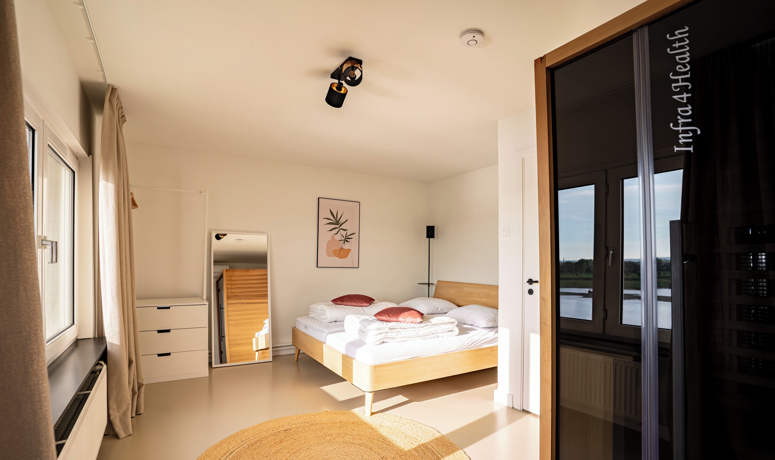 A bedroom with a bed, a bedside table, a mirror, a wall-mounted light, and a window with a view.