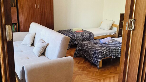A room with a sofa, two beds with patterned bedding, a wooden wardrobe, and a herringbone floor.