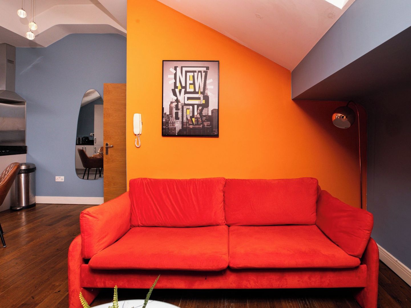 A room with a red sofa, a cityscape poster, a mirror, and a kitchen area.