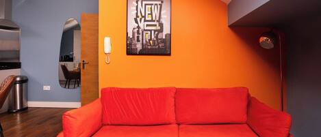 A room with a red sofa, a cityscape poster, a mirror, and a kitchen area.