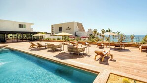 Un espace piscine avec des chaises longues et des parasols, un bâtiment portant l’inscription « Grand Velas », et une vue sur l’océan.