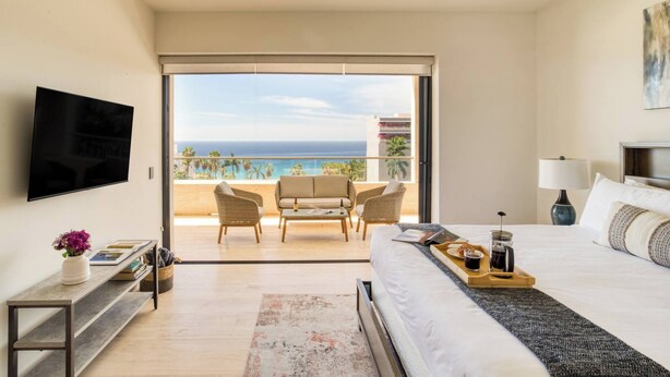 Une chambre d’hôtel comprenant un lit, une télévision, un balcon avec vue sur l’océan et un coin salon avec des chaises et une table.