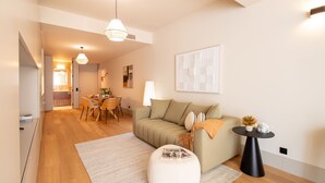 Una sala de estar moderna con un sofá, una mesa de comedor y sillas. Hay una mesa de centro y una lámpara. El piso es de madera y las paredes son blancas.
